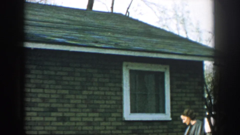 1957 Wisconsin frame with a woman partly visible at the right edge of a brick house wall and white-framed window.