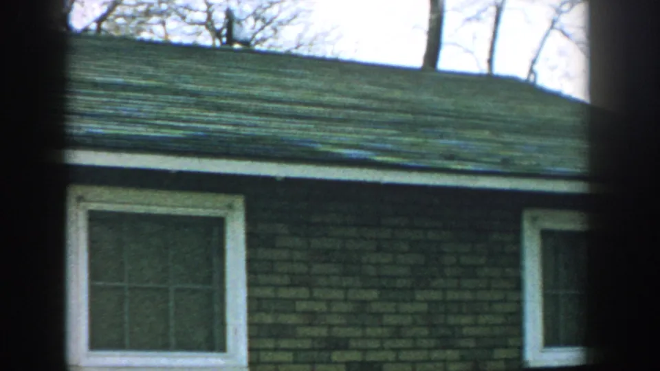 1957 Wisconsin home-movie frame focused on the brick house side, white-trimmed windows, roof shingles, and bare trees above.