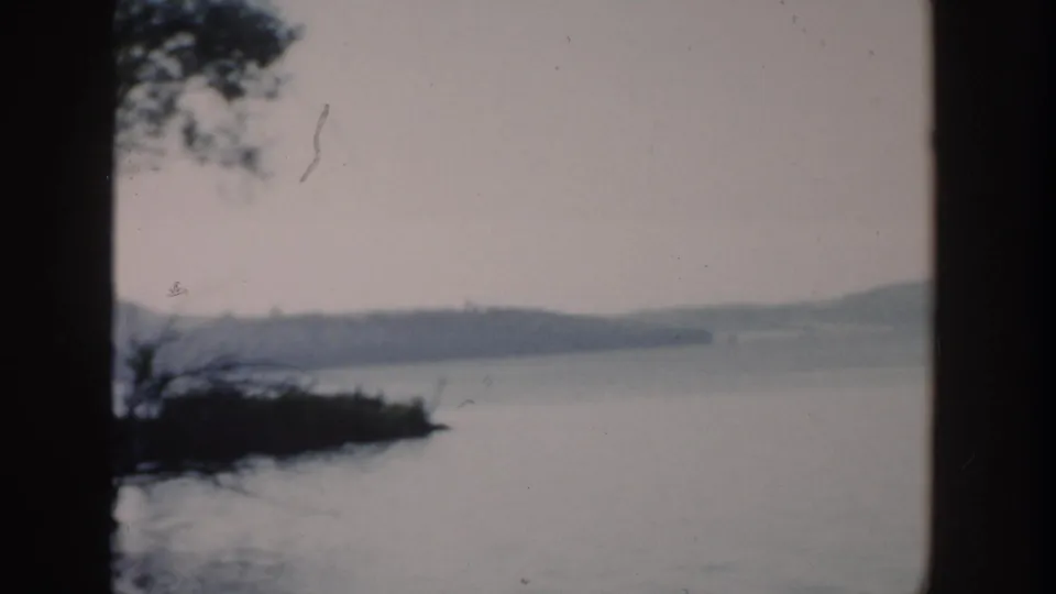 1957 Wisconsin lake scene with shoreline trees in silhouette, still water, hazy hills, and dark frame edges.
