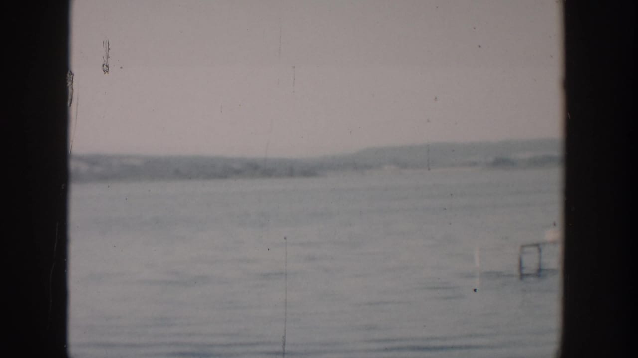 1957 Wisconsin home-movie view across calm water toward a hazy shore, with faint dock-like structures briefly visible.