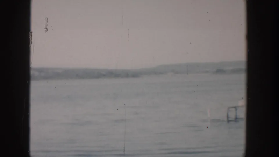 1957 Wisconsin home-movie view across calm water toward a hazy shore, with faint dock-like structures briefly visible.