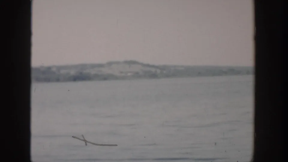 1957 Wisconsin 8mm frame showing rippled water in the foreground and a hazy low shoreline across the distance.
