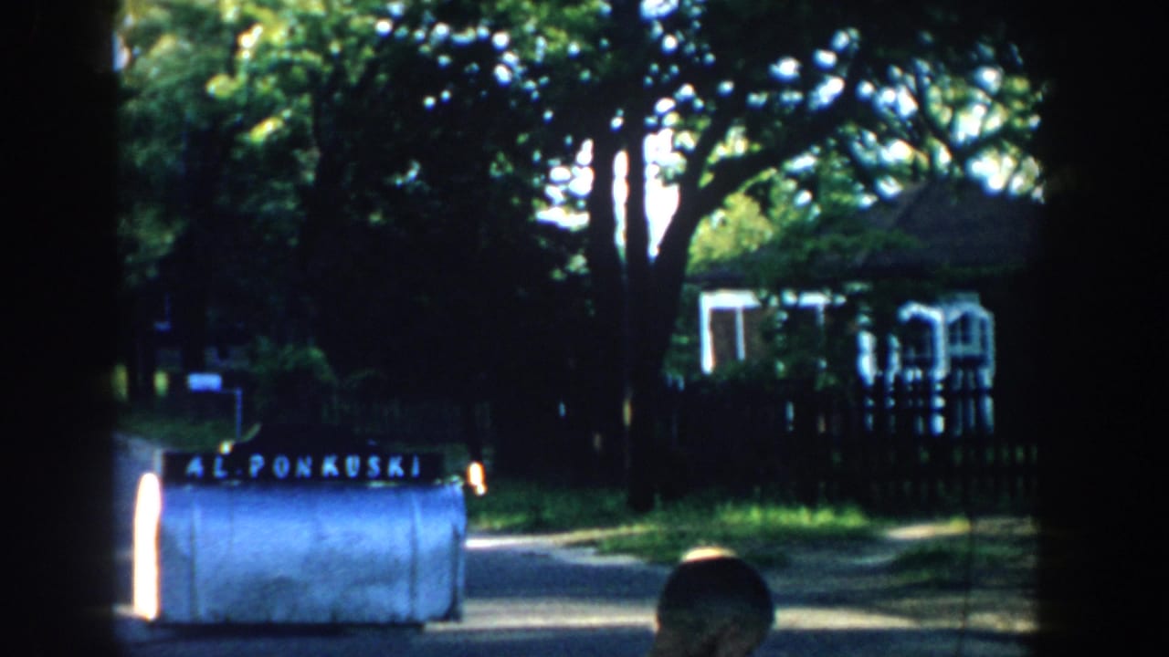 Dark-edged 8mm frame of a suburban street with a white machine and trees in the background.