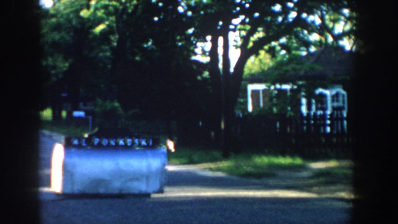 White machine on a residential driveway or street edge with trees, fence, and house behind it.