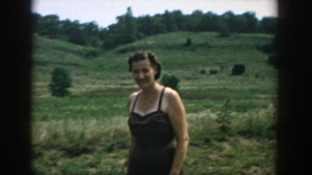 Woman in a dark swimsuit standing in a grassy field with rolling hills behind her.