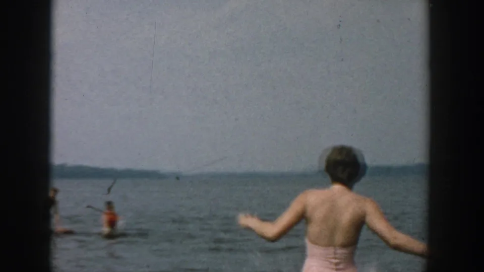 1957 Wisconsin lakeside frame showing a woman in a pink swimsuit reaching out near swimmers in the water.