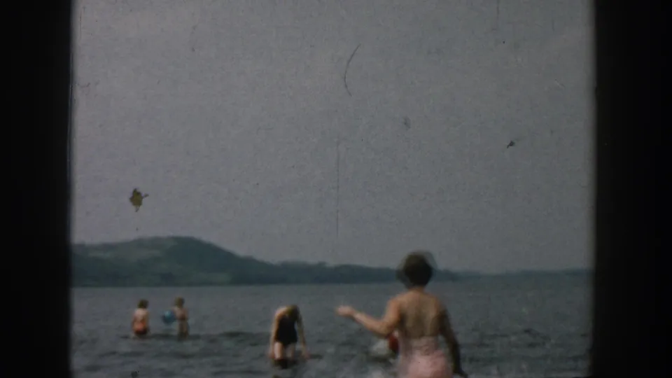 1957 Wisconsin home-movie frame with swimmers in a lake, a woman at shore, and a bird above the water.