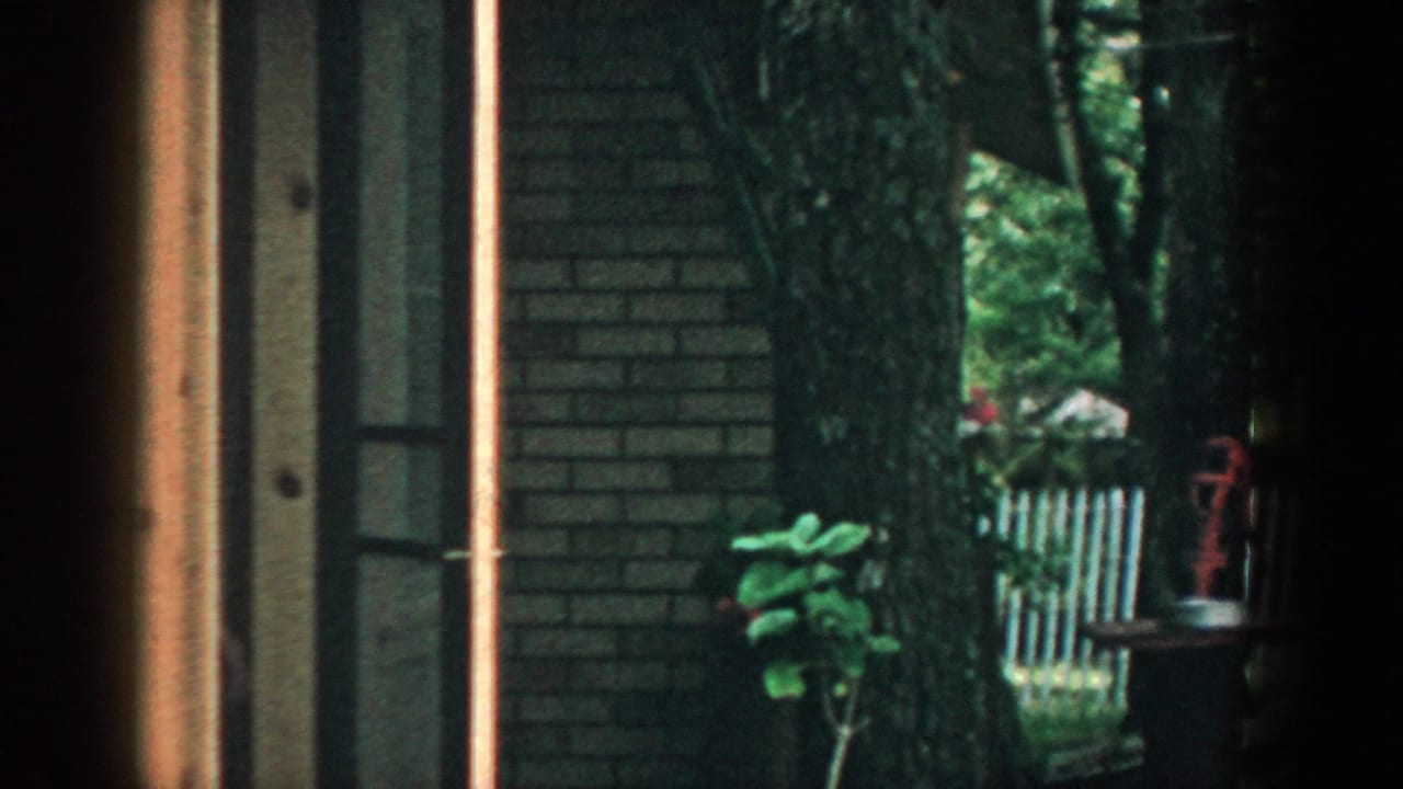 1957 Wisconsin 8mm frame looking through a window toward a wooded backyard with a brick house edge and green covered structure.