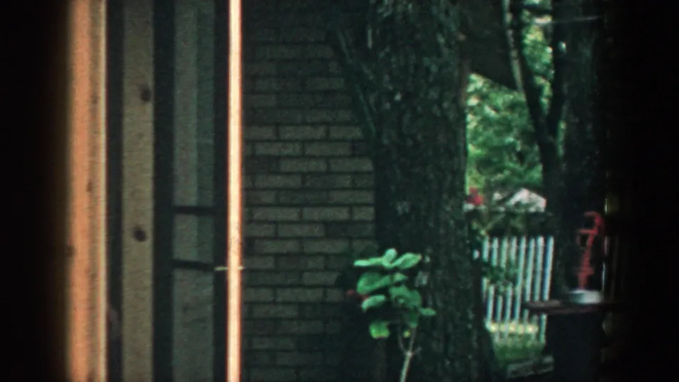 1957 Wisconsin 8mm frame looking through a window toward a wooded backyard with a brick house edge and green covered structure.