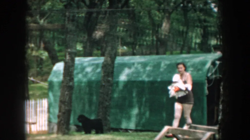 1957 Wisconsin home-movie frame of a woman in a dark swimsuit walking through the backyard with a white towel and small black dog nearby.