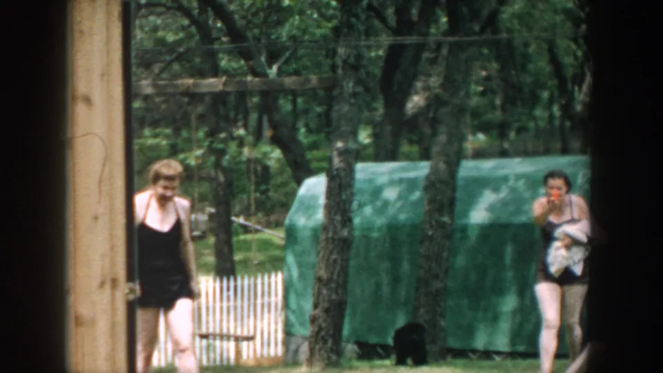 1957 Wisconsin 8mm frame showing the woman with a towel near a white fence, trees, green covered structure, and small black dog.