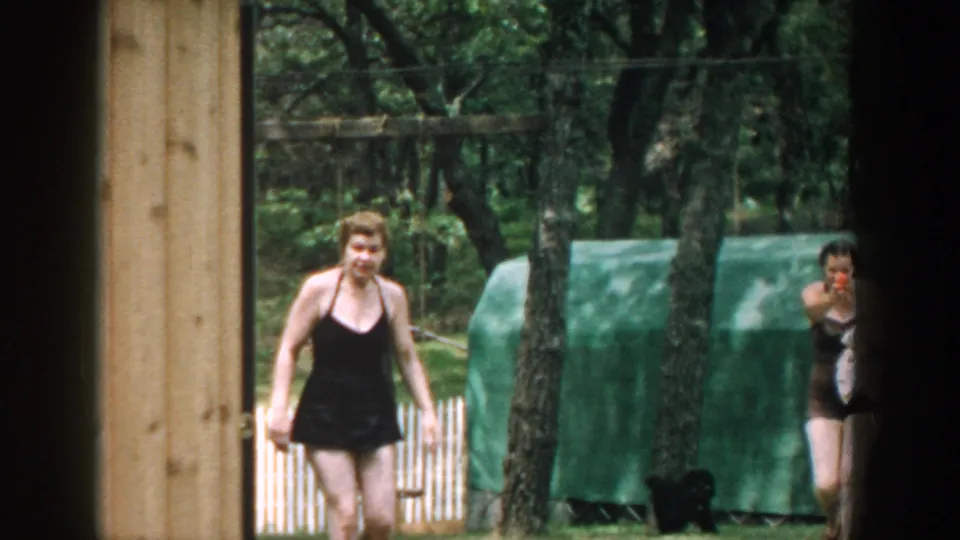1957 Wisconsin home-movie frame showing a woman in a black one-piece bathing suit walking through a wooded yard near a small building.