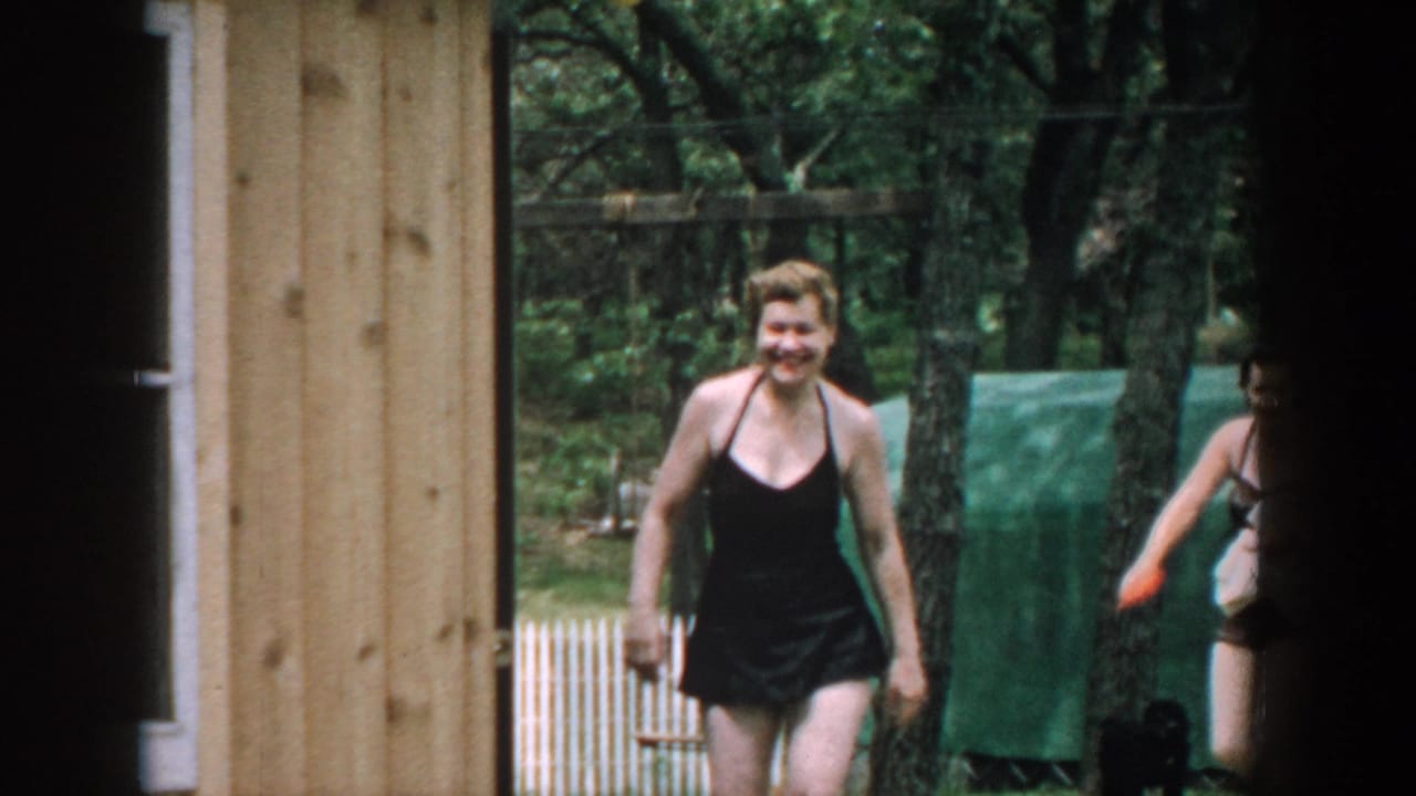 Smiling woman in a black bathing suit walking past a wooden building outdoors.