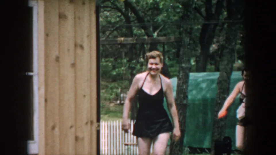 1957 Wisconsin frame with a smiling woman in a black one-piece bathing suit walking beside a wooden building in a tree-lined yard.