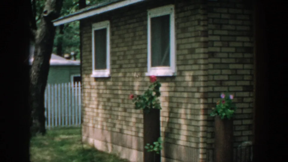 1957 Wisconsin home-movie frame showing a small brick building exterior with windows, flowers, trees, and a white fence nearby.