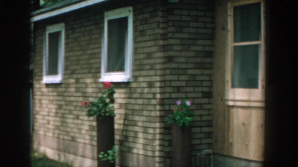 1957 Wisconsin frame showing a small brick building, wooden door, windows, and flower planters in a shaded outdoor setting.
