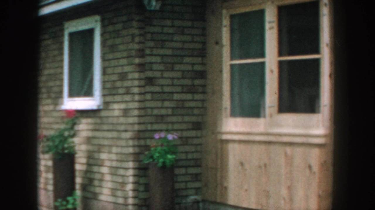 Wood and brick cabin exterior with windows, grass, trees, and two women partly visible near the right edge.