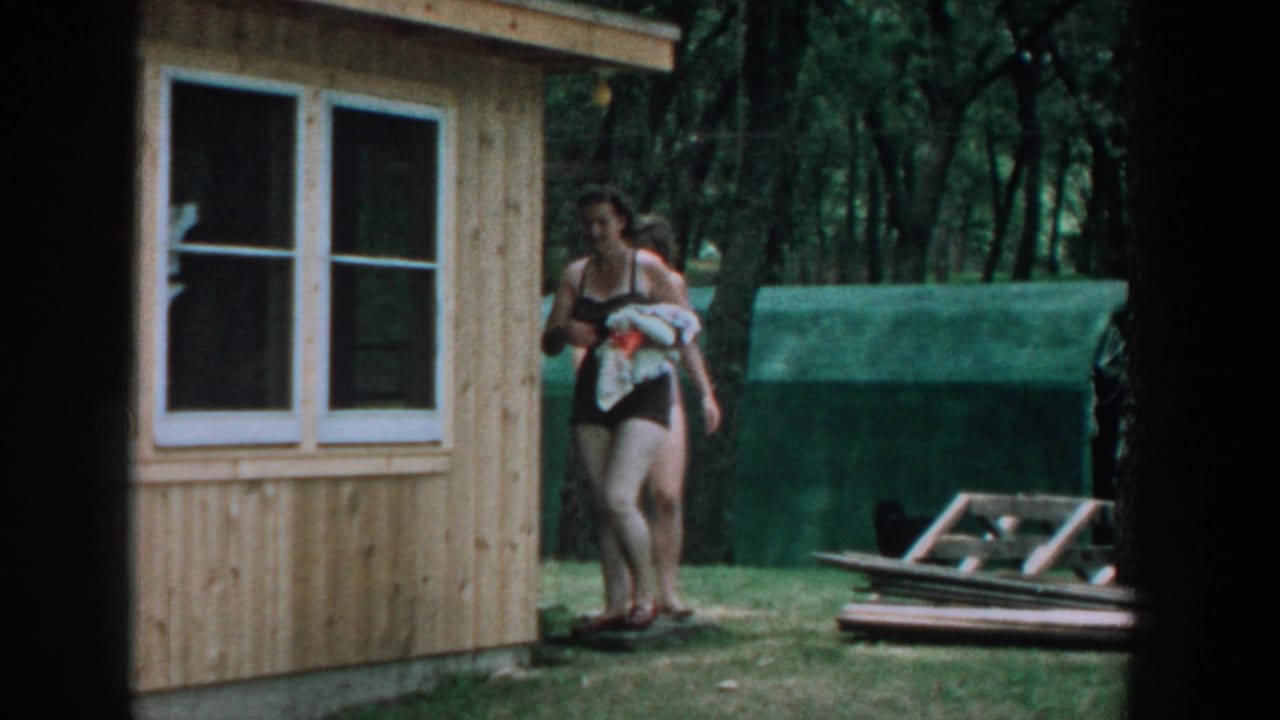 1957 Wisconsin home-movie frame of two women in bathing suits walking by a wooden cabin in a wooded summer setting.