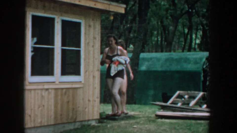 1957 Wisconsin home-movie frame of two women in bathing suits walking by a wooden cabin in a wooded summer setting.