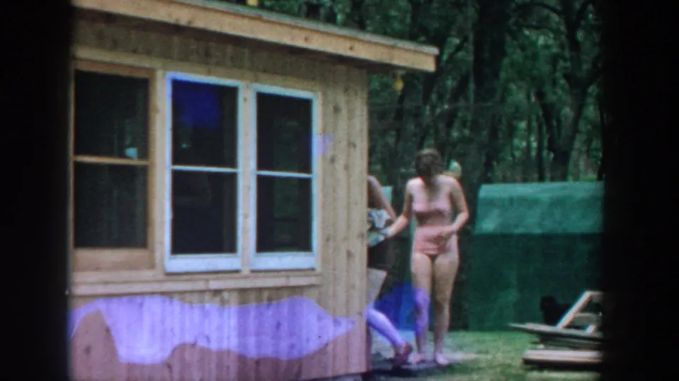 1957 Wisconsin 8mm frame showing two women in swimsuits beside a wood-sided cabin, with a towel and wooded grounds visible.