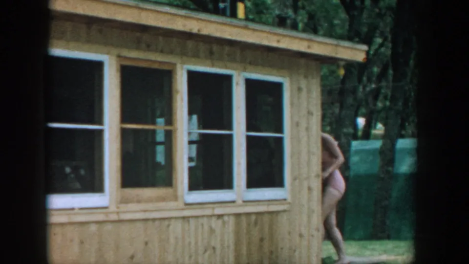1957 Wisconsin home-movie frame of a wood-sided cabin and white windows, with a woman in a bathing suit partly visible at right.