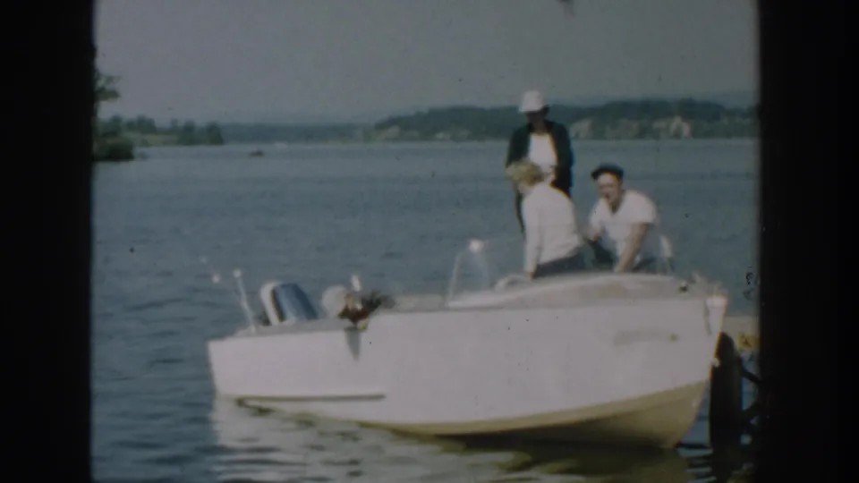 A 1957 Wisconsin home-movie frame shows three adults on a white motorboat beside the dock before departure.