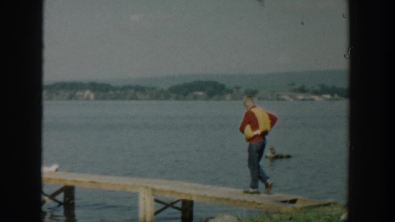 Man in a yellow life vest stands on a wooden dock beside calm water.