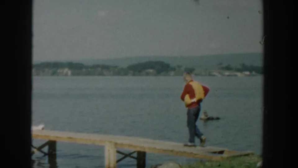 1957 Wisconsin home-movie frame showing a man in a yellow life vest on a wooden dock with lake or river water and distant shoreline behind him.