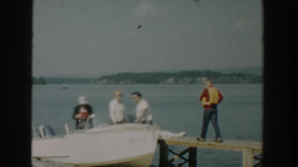 Adults gather in and around a small white motorboat beside a wooden dock, with a man in a yellow life vest standing near the edge in the 1957 Wisconsin footage.