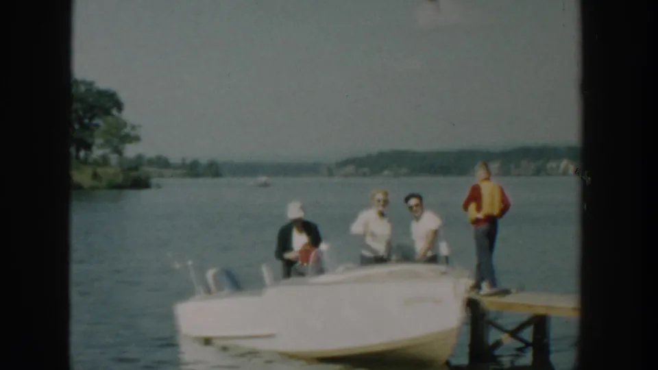 The boat is beside the dock with several adults visible, including a woman in a white hat, while a man in a yellow life vest stands at the dock edge.