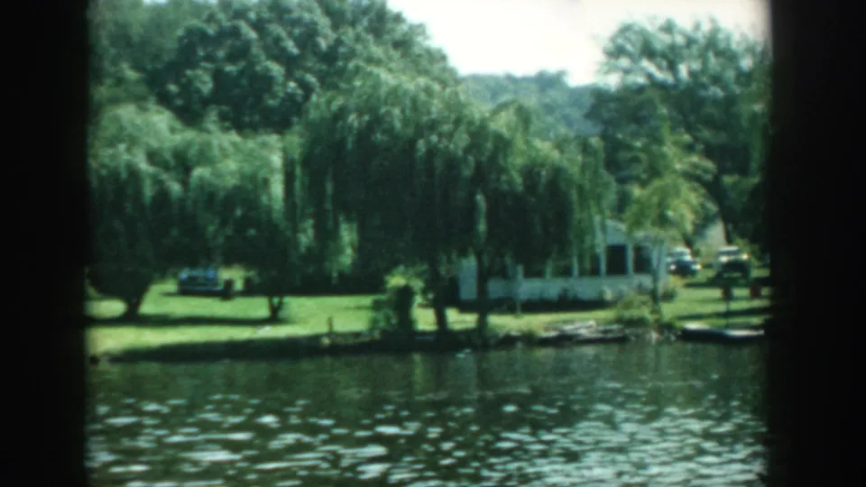 1957 Wisconsin frame with rippling lake water in front of a green lawn, weeping willows, a white house, and a road or driveway beyond.