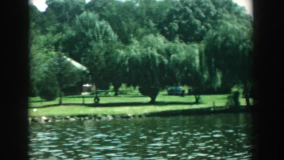 1957 Wisconsin 8mm frame looking across water to a willow-shaded lawn, parked car, shoreline rocks, and small distant figures.