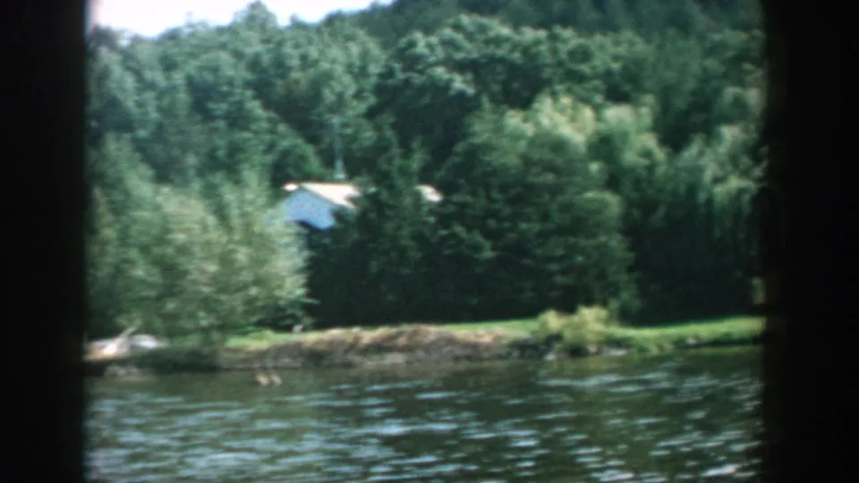 1957 Wisconsin 8mm frame showing lake water, a leafy shoreline, dense trees, and a small pale-roofed structure partly obscured.