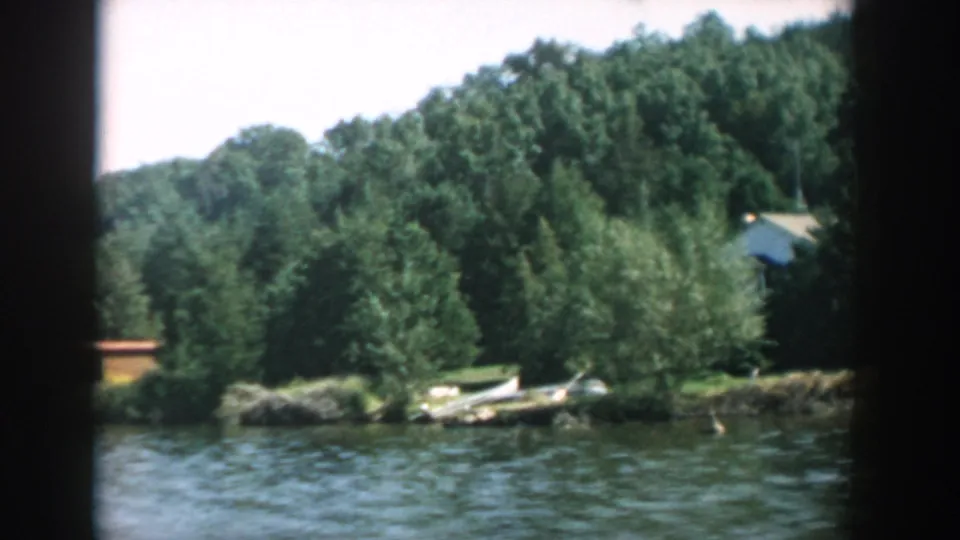 1957 Wisconsin lake view with rippled water, a wooded bank, a small white boat on shore, and a structure partly visible through trees.