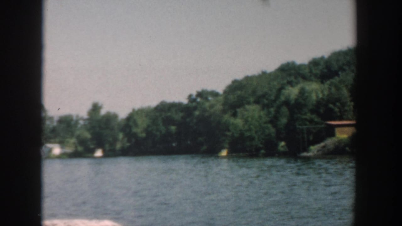 1957 Wisconsin home-movie frame of a broad river view with tree-lined banks and distant waterside structures.