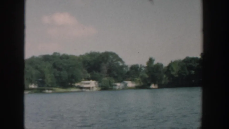 1957 Wisconsin 8mm frame showing a wide river, low shoreline trees, and a pale building near the waterline.