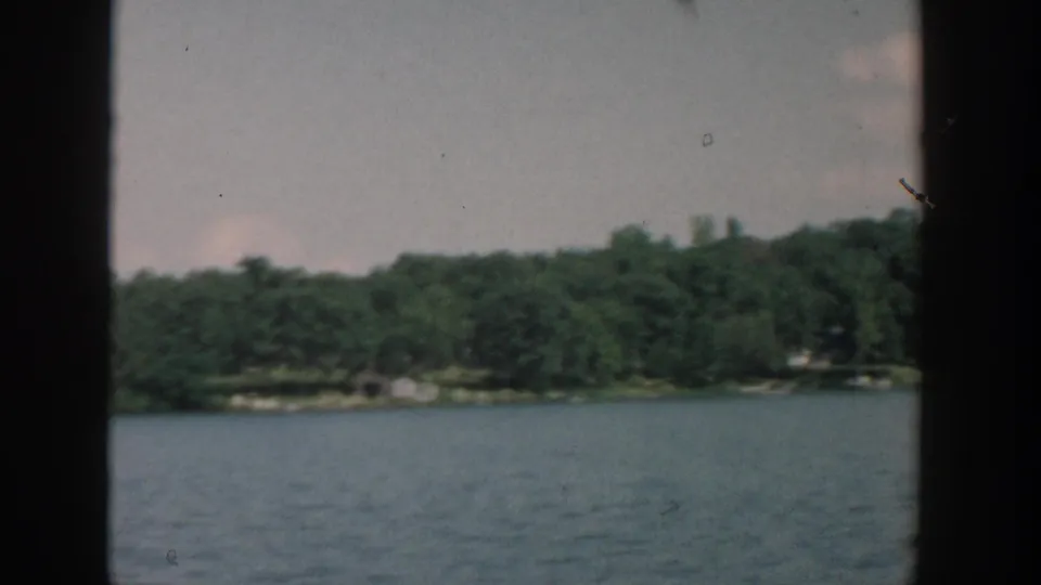 1957 Wisconsin home-movie frame of calm water and a wooded bank with faint distant structures beyond the trees.