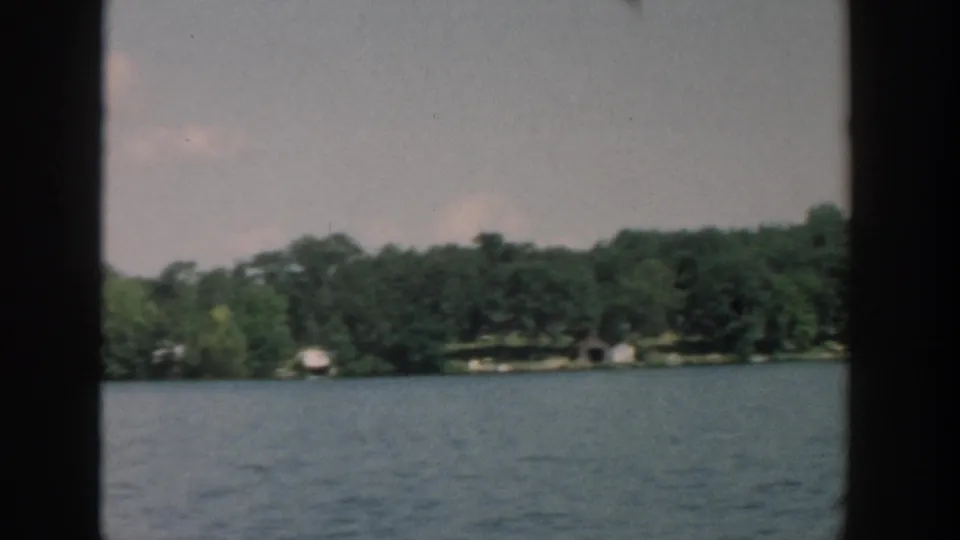 1957 Wisconsin 8mm frame showing lake water, wooded shoreline, and small distant lakeside structures from a boat viewpoint.
