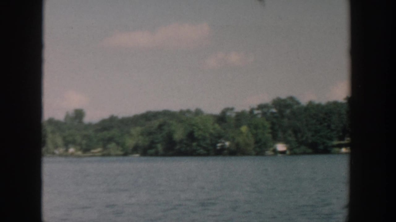 1957 Wisconsin lake view with open water, dense trees, and distant shore structures framed by dark vertical edges.