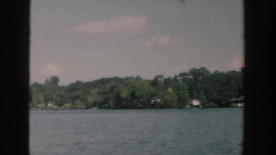 1957 Wisconsin lake view with open water, dense trees, and distant shore structures framed by dark vertical edges.