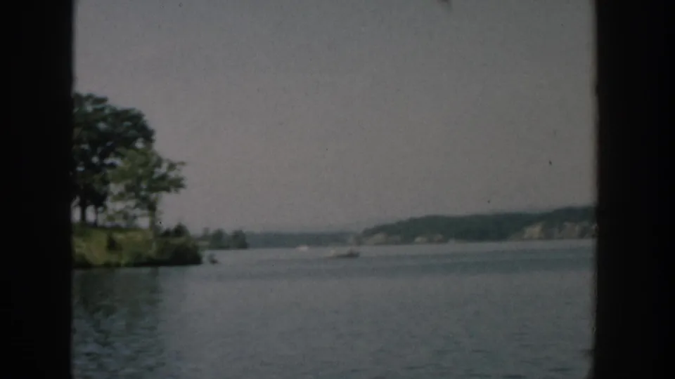 1957 Wisconsin 8mm view of a small wooded island across calm water, seen from a moving boat with distant shoreline beyond.