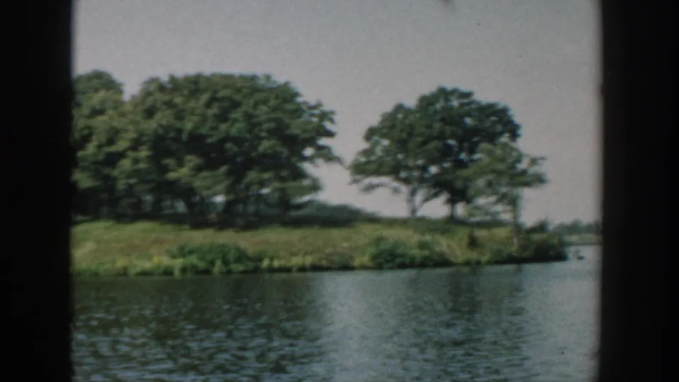 1957 Wisconsin 8mm boat view passing the island edge, with rippled water, clustered trees, and a soft pale sky.