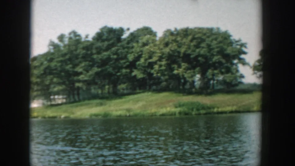 1957 Wisconsin lake scene with a closer view of dense trees on a small island and rippled water in the foreground.