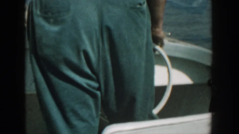 1957 Wisconsin home-movie frame showing a close view of an adult's green trousers near a white motorboat railing with lake water behind.