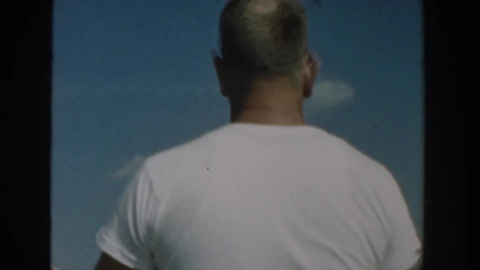 Back view of an adult man in a white t-shirt filmed from a motorboat, with blue sky and clouds in the 1957 Wisconsin lake scene.