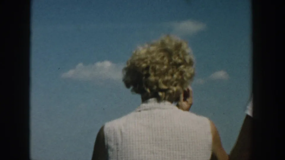 1957 Wisconsin home-movie frame showing a woman from behind on a boat, with blue sky, clouds, and a narrow glimpse of another adult.