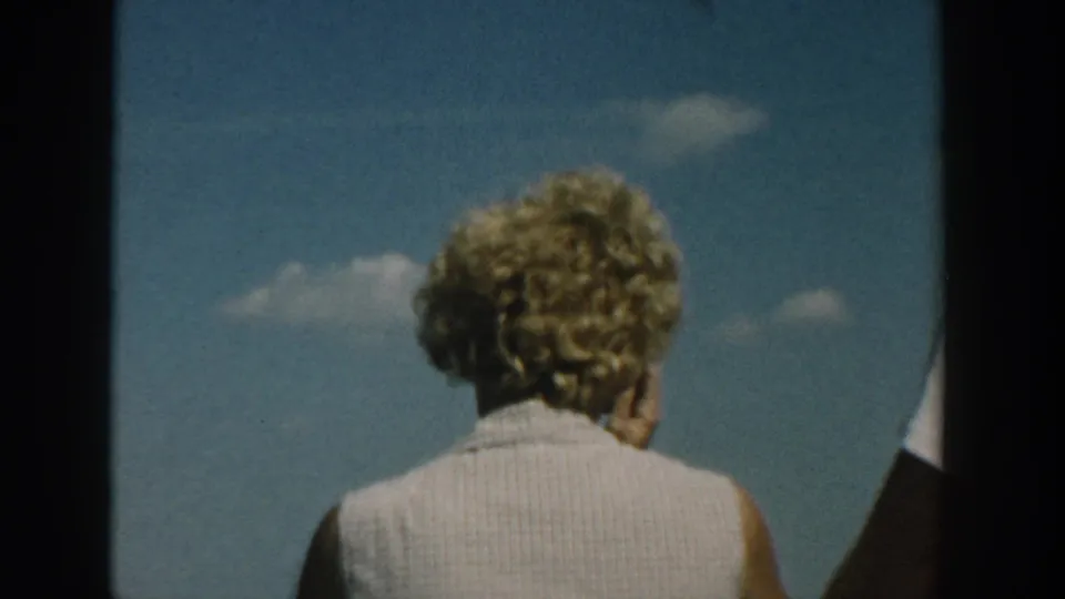 1957 Wisconsin 8mm frame showing a blonde woman from behind in a sleeveless light top under a blue sky.