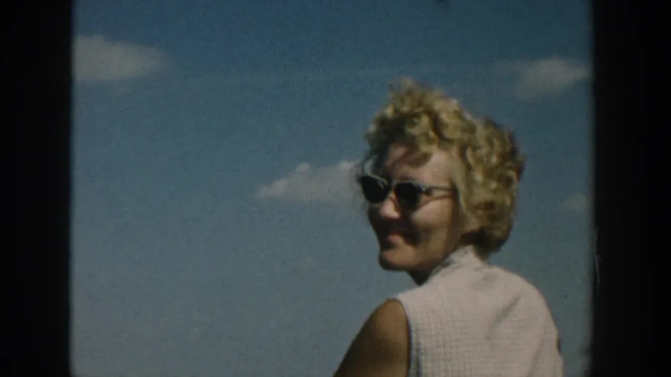 1957 Wisconsin home-movie frame of a blonde woman in dark sunglasses turning to smile outdoors.