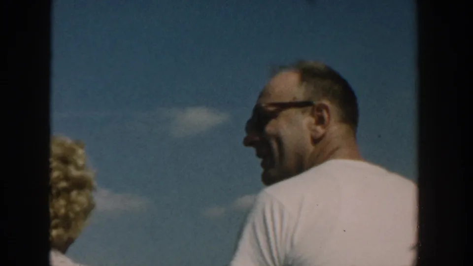 1957 Wisconsin 8mm frame with a man in sunglasses and a white T-shirt beside the blonde woman outdoors.