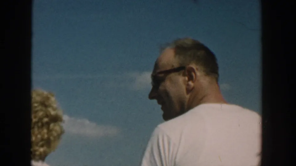 1957 Wisconsin home-movie frame showing a man in glasses beside the partly visible blonde woman beneath a blue sky.